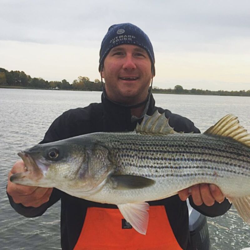 The image shows a man holding a striped bass fish. He is wearing a blue beanie, a black jacket, and an orange bib. The background shows a body of water and a cloudy sky. The man is smiling, suggesting he is happy with his catch.